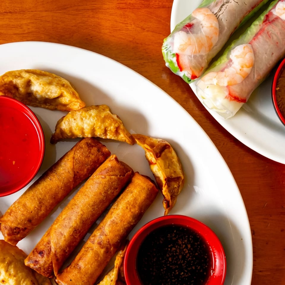 Fried spring rolls, dumplings, fresh shrimp rolls, and three dipping sauces on white plates.