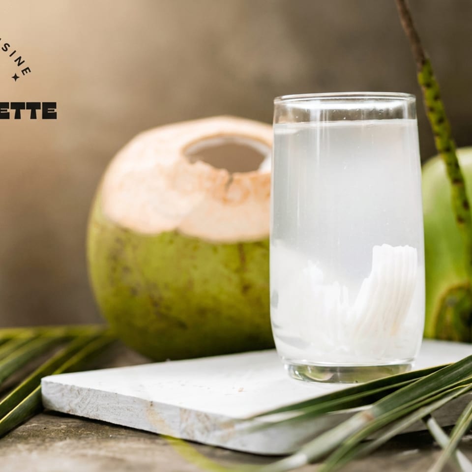 Glass of fresh coconut water with meat, surrounded by whole and opened coconuts and palm leaves.